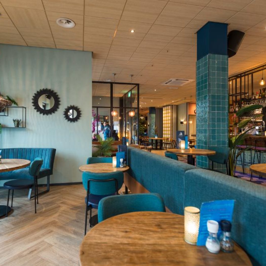 Modern restaurant met blauwgroene zithoeken, ronde houten tafels en een betegelde bar.