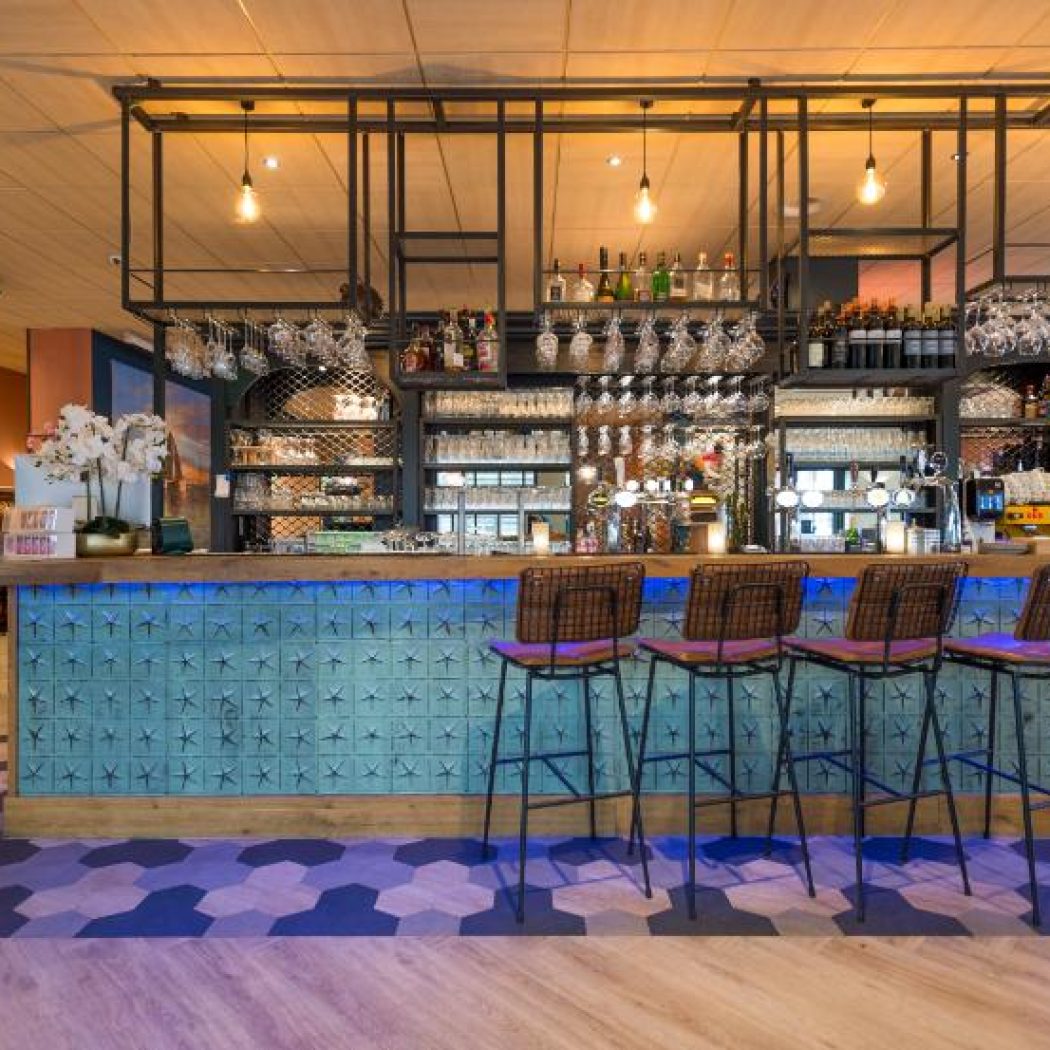 Moderne bar in restaurant met turquoise sterrentegels, blauwe LED-verlichting en vier barkrukken met paarse zittingen.