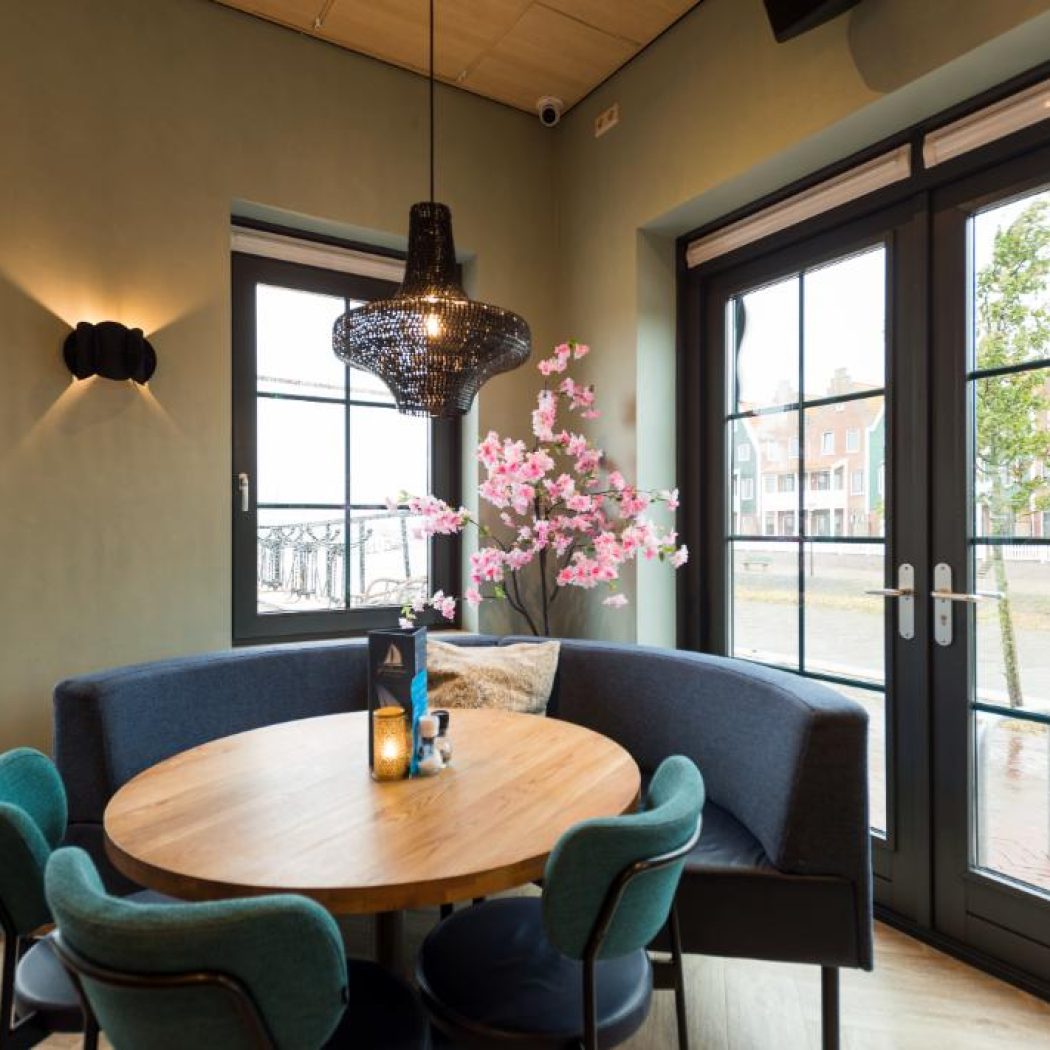 Sfeervolle restaurant eethoek met ronde houten tafel, donkerblauwe bank en turkooizen stoelen bij ramen met uitzicht.