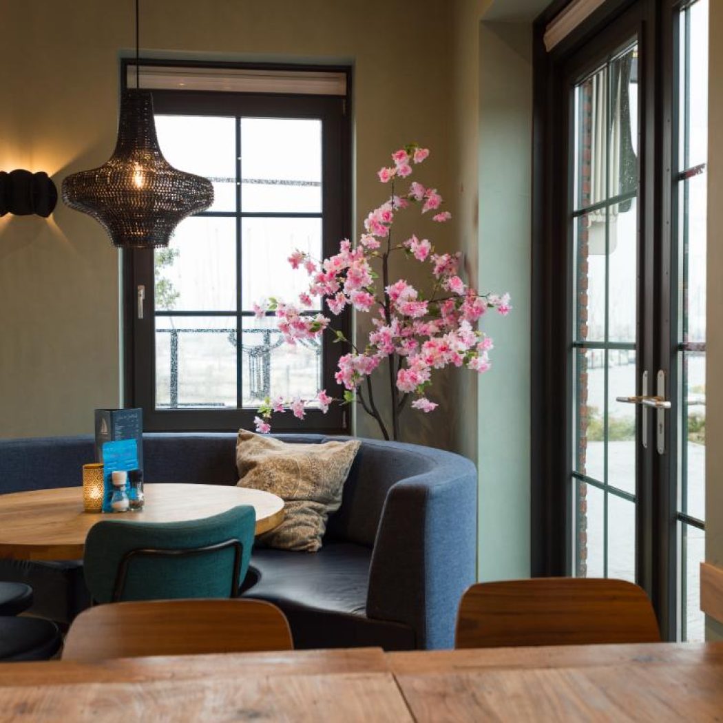 Sfeervol restaurant interieur met blauwe booth, ronde houten tafel, teal stoelen en roze bloesemboom bij grote ramen.