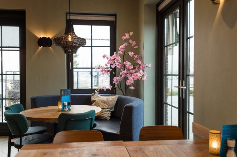 Sfeervol restaurant interieur met blauwe booth, ronde houten tafel, teal stoelen en roze bloesemboom bij grote ramen.