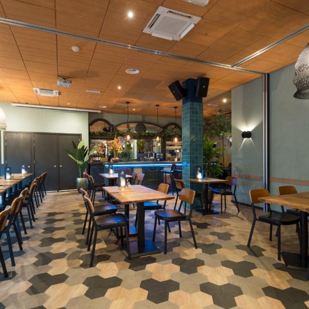 Sfeervol restaurantinterieur met houten tafels, donkere stoelen, een bar met blauwe verlichting en een hexagonale vloer.