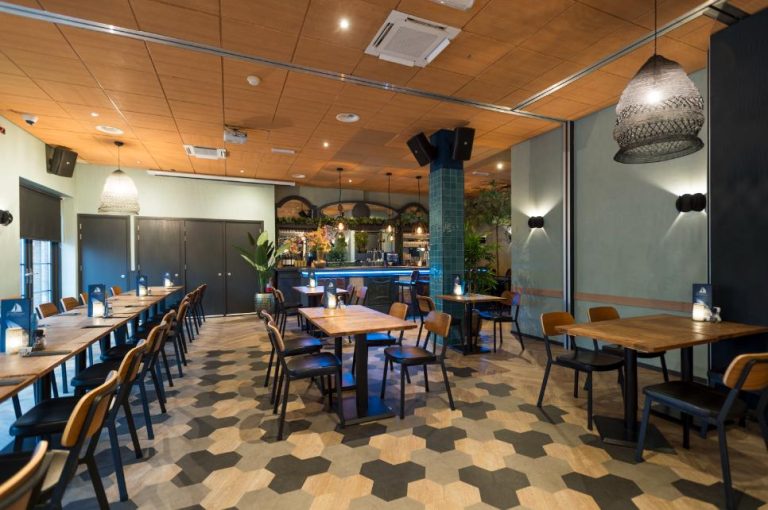 Sfeervol restaurantinterieur met houten tafels, donkere stoelen, een bar met blauwe verlichting en een hexagonale vloer.