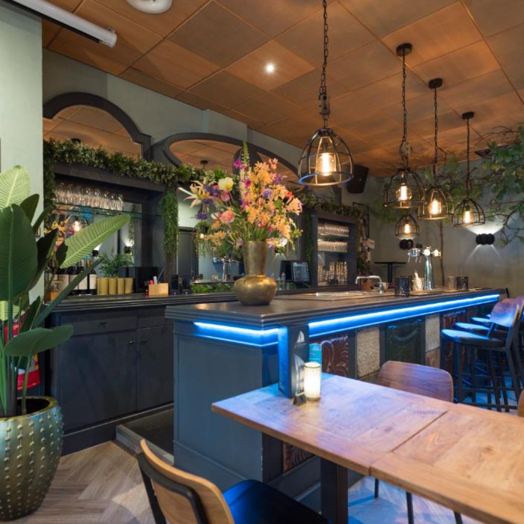 Modern restaurant interieur met bar met blauwe LED-verlichting, barstoelen, bloemstuk en industriële hanglampen.