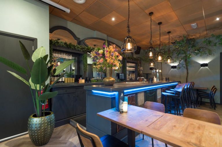 Modern restaurant interieur met bar met blauwe LED-verlichting, barstoelen, bloemstuk en industriële hanglampen.