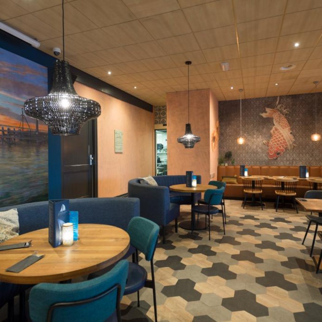 Stijlvol restaurant met blauwe en bruine banken, houten tafels, muurschilderingen van een zeilboot en koi, en zeshoekige vloer.