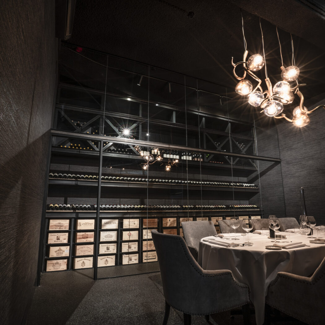 Luxe donkere eetzaal met wijnrek vol flessen en kisten, gedekte tafel en moderne kroonluchter.