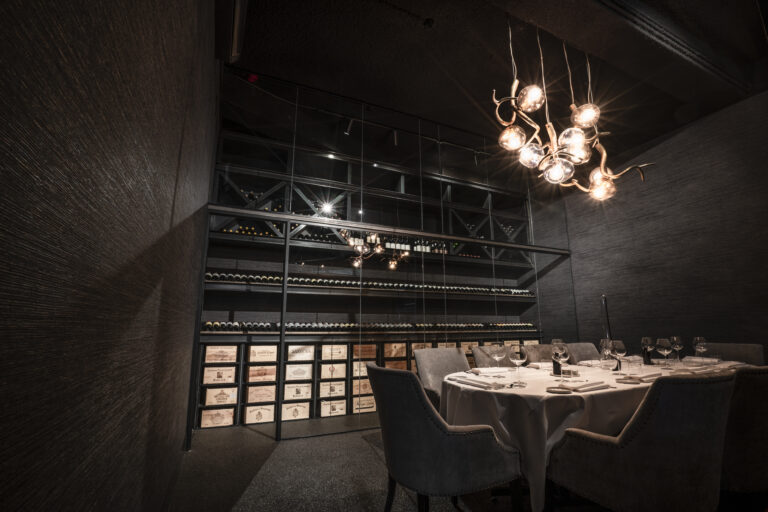 Luxe donkere eetzaal met wijnrek vol flessen en kisten, gedekte tafel en moderne kroonluchter.