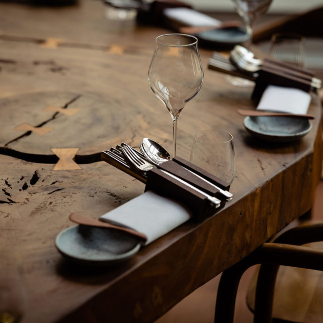 Restauranttafel met glazen, bestek in een houten houder en een servet op een robuust houten blad met inlegwerk.