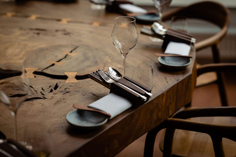 Restauranttafel met glazen, bestek in een houten houder en een servet op een robuust houten blad met inlegwerk.