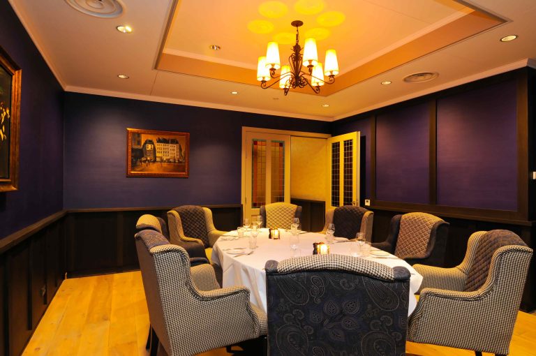 Sfeervolle privé-eetkamer met ronde tafel gedekt voor diner, diverse fauteuils en warme kroonluchterverlichting.
