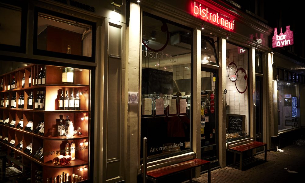 Avondzicht op bistrot neuf met rode neonreclame, aangrenzende wijnwinkel met flessen en bar à vin neonbord.