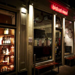 Avondzicht op bistrot neuf met rode neonreclame, aangrenzende wijnwinkel met flessen en bar à vin neonbord.