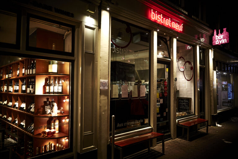 Avondzicht op bistrot neuf met rode neonreclame, aangrenzende wijnwinkel met flessen en bar à vin neonbord.