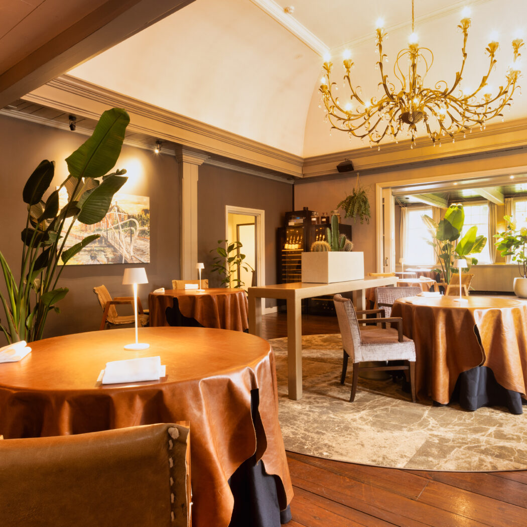 Warm verlicht restaurant met leren tafels, gouden kroonluchter en weelderige planten.