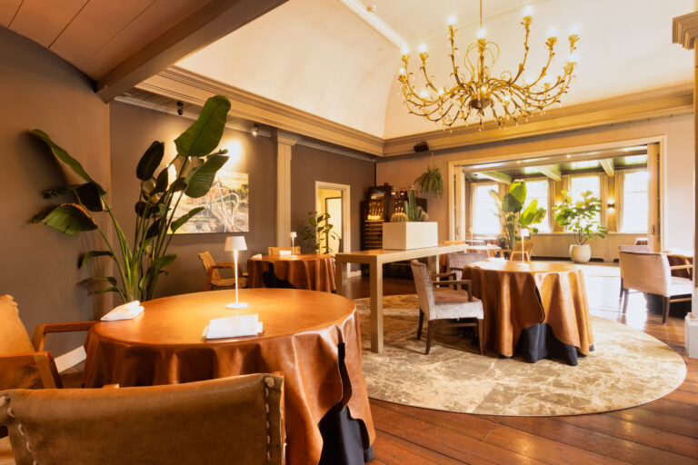 Warm verlicht restaurant met leren tafels, gouden kroonluchter en weelderige planten.