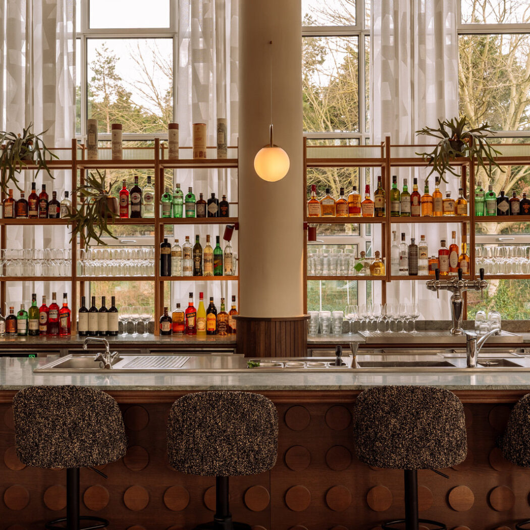 Lichte moderne bar met marmeren balie, houten schappen vol drankflessen en gestoffeerde barkrukken voor grote ramen.