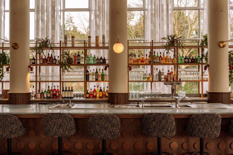 Lichte moderne bar met marmeren balie, houten schappen vol drankflessen en gestoffeerde barkrukken voor grote ramen.