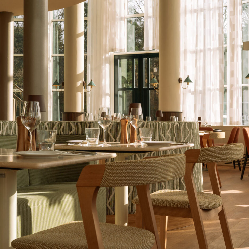 Elegant restaurantinterieur met gedekte houten tafels, groene zithoeken en grote ramen met zonlicht.