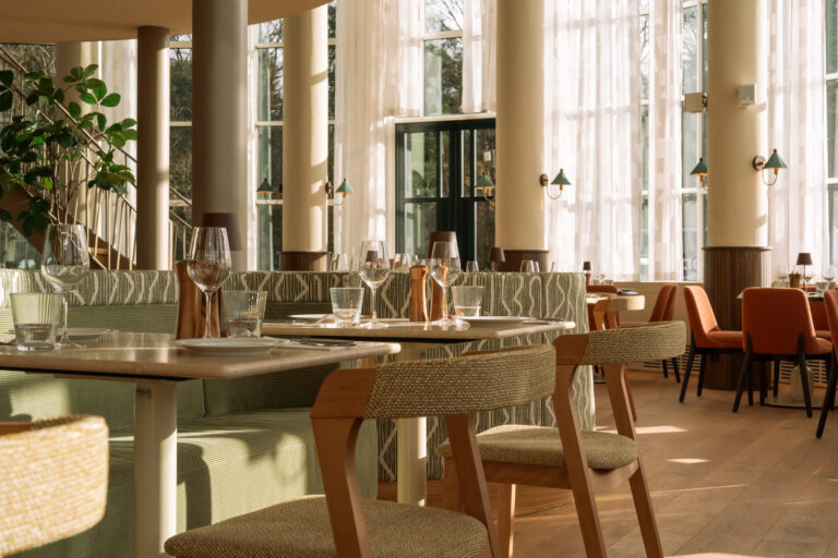 Elegant restaurantinterieur met gedekte houten tafels, groene zithoeken en grote ramen met zonlicht.