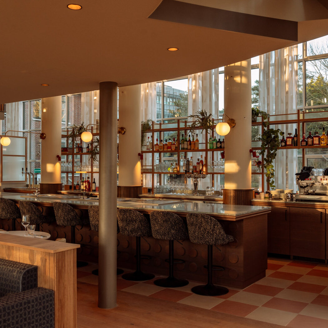Modern en warm verlicht bar interieur met balie, gevulde drankplanken en grote ramen.