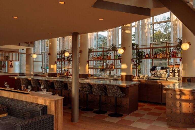 Modern en warm verlicht bar interieur met balie, gevulde drankplanken en grote ramen.