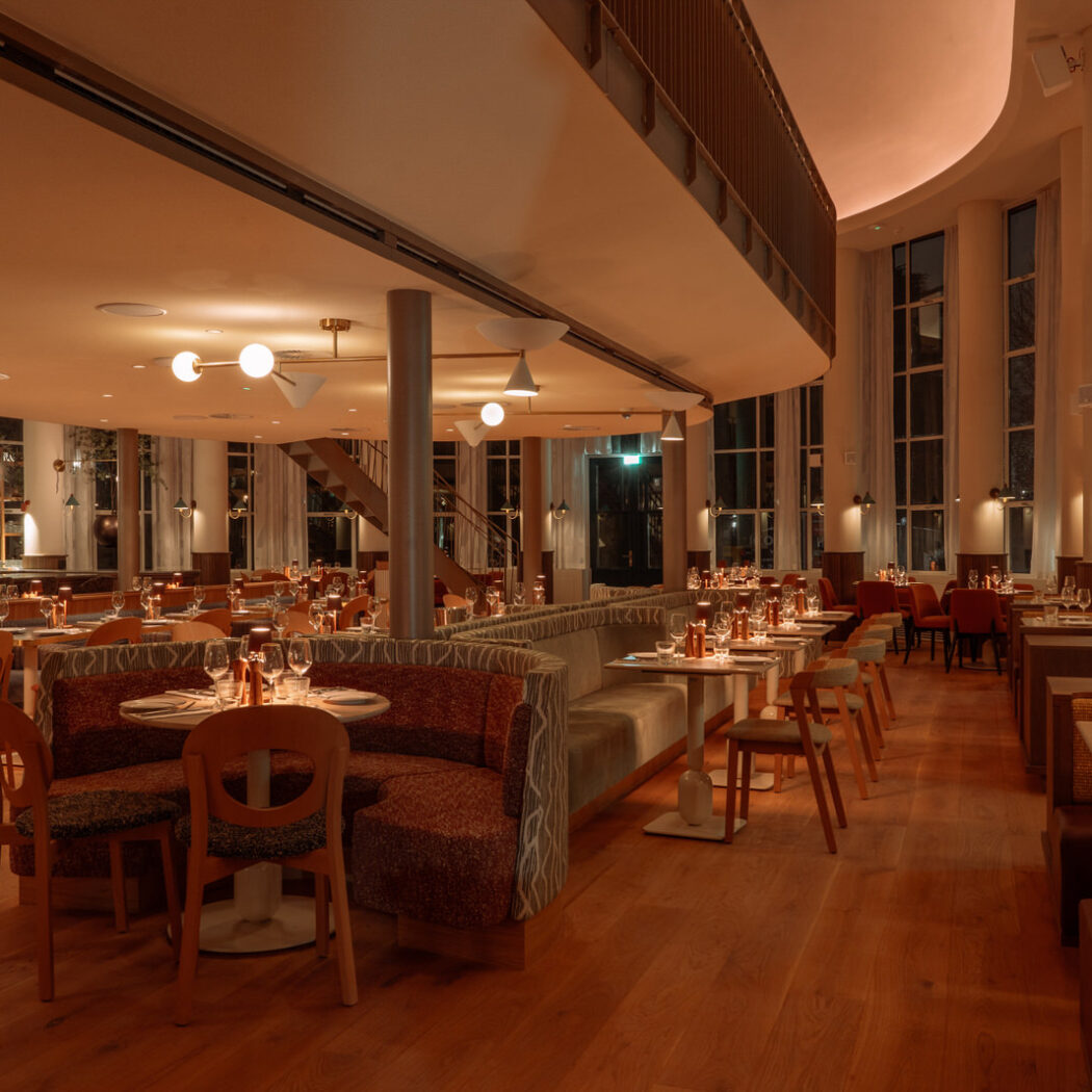Ruim restaurant met gebogen gestoffeerde banken, houten stoelen en gedekte tafels, sfeervol warm verlicht.
