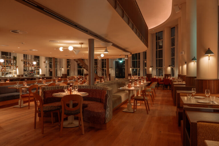 Ruim restaurant met gebogen gestoffeerde banken, houten stoelen en gedekte tafels, sfeervol warm verlicht.
