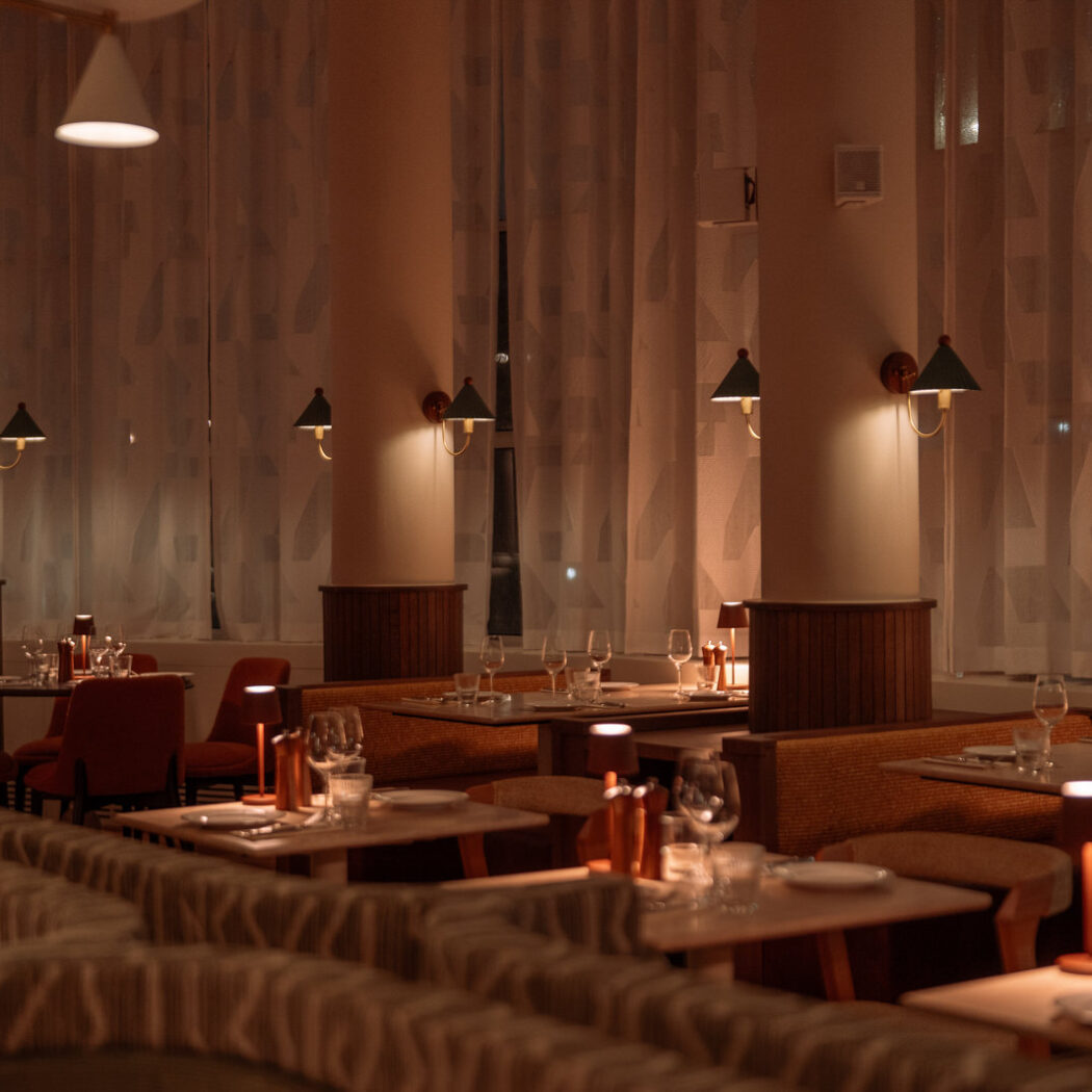 Sfeervol dinerrestaurant met gedekte tafels, banken en warme verlichting bij hoge ramen.