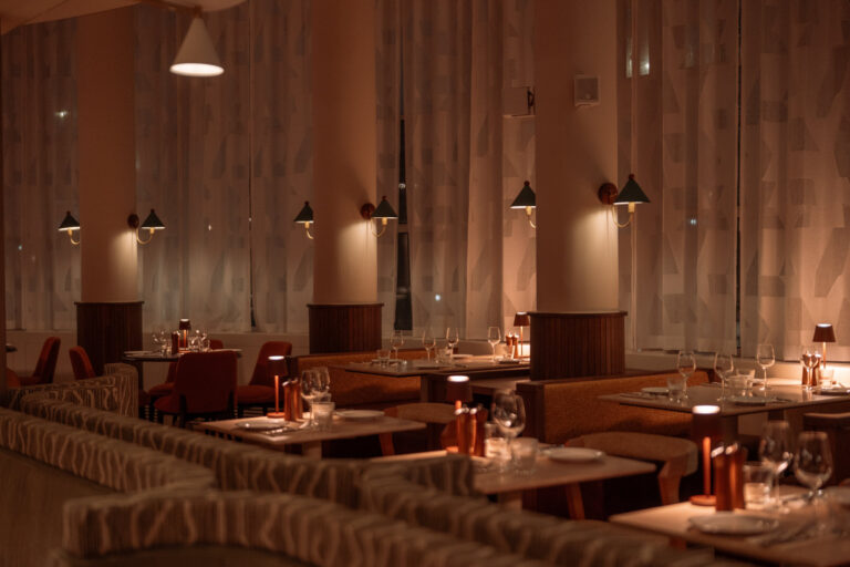 Sfeervol dinerrestaurant met gedekte tafels, banken en warme verlichting bij hoge ramen.