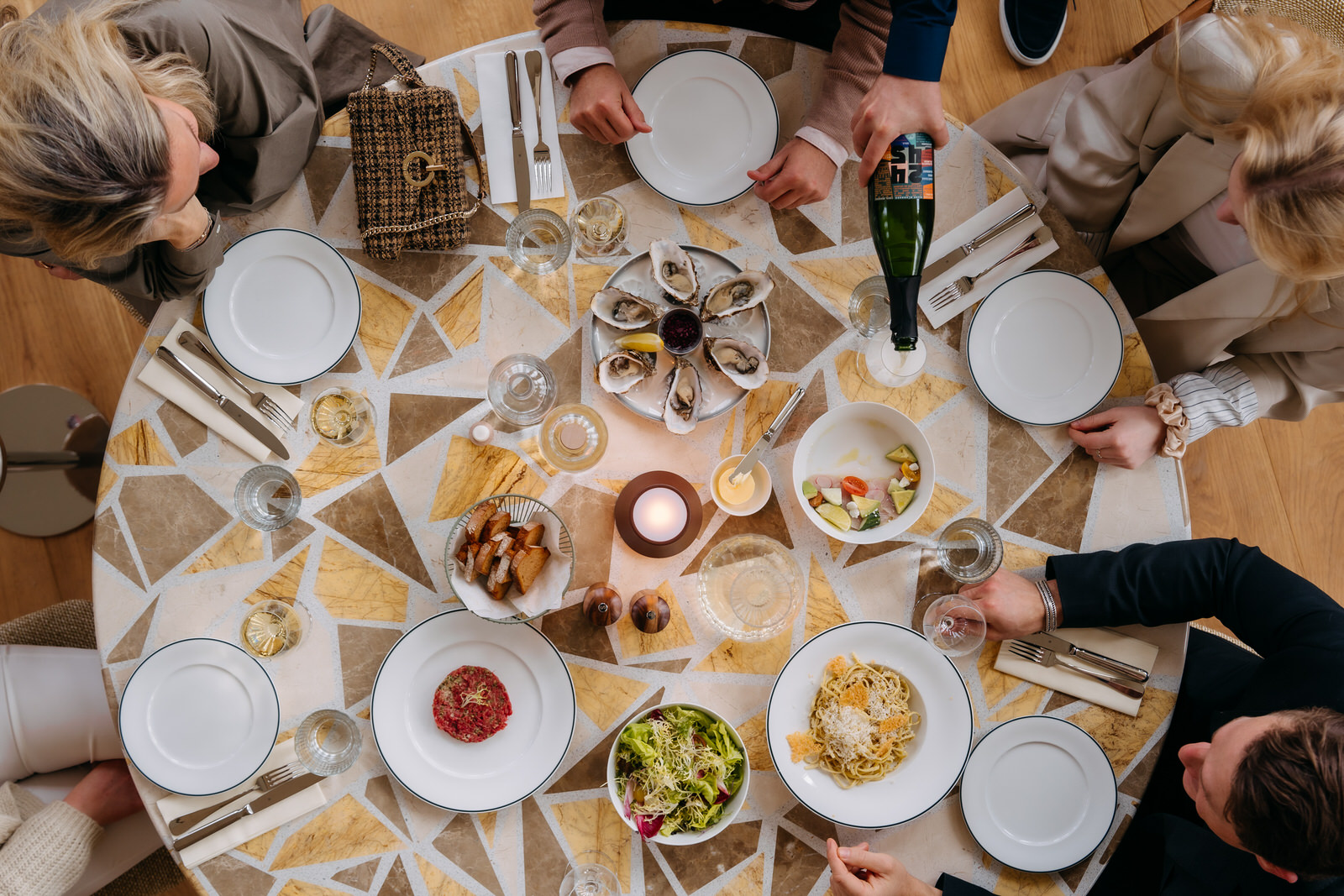 Sfeervol diner aan ronde mozaïektafel vol oesters, pasta, salade en wijn, waar gasten van de maaltijd genieten.