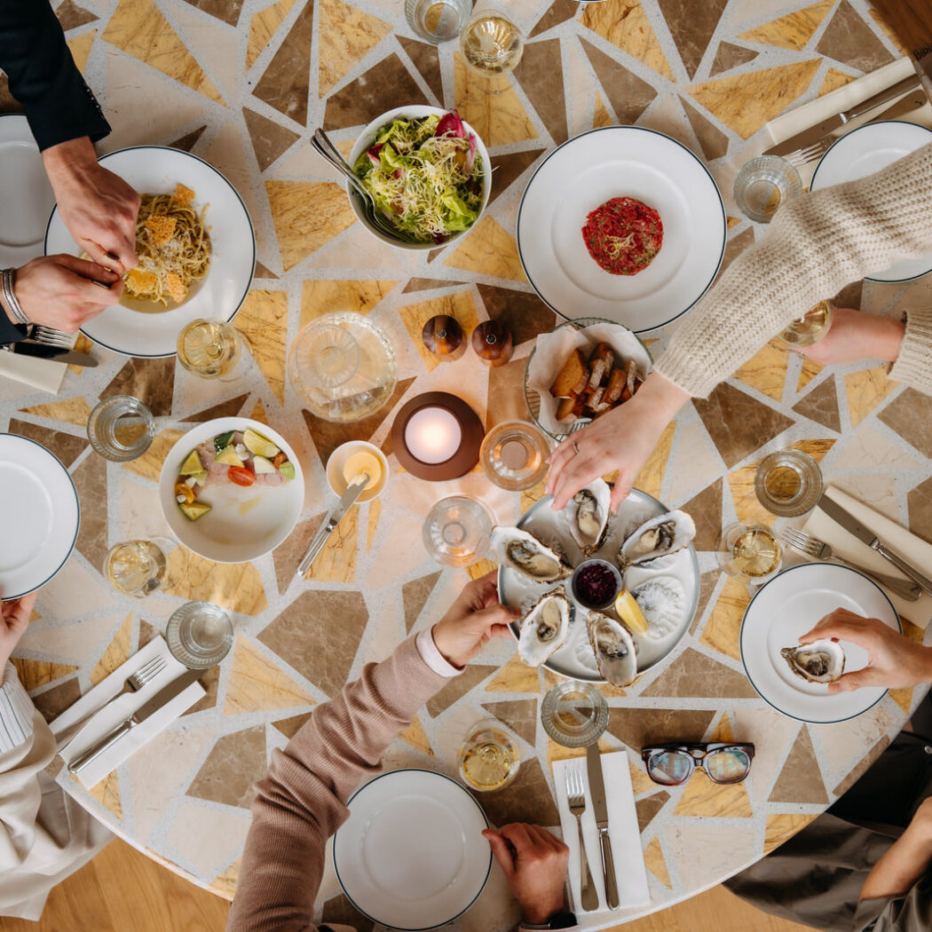 Mensen delen diverse gerechten zoals oesters, pasta en tartaar aan een mozaïektafel met wijn en kaarslicht.