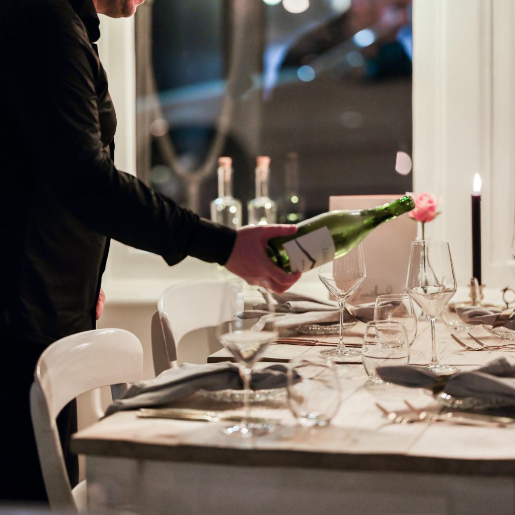 Man in zwart overhemd schenkt wijn uit een groene fles aan een elegant gedekte tafel.
