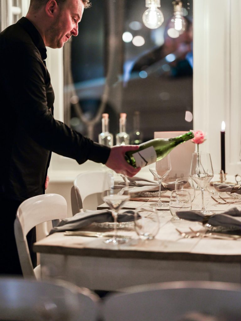 Man in zwart overhemd schenkt wijn uit een groene fles aan een elegant gedekte tafel.