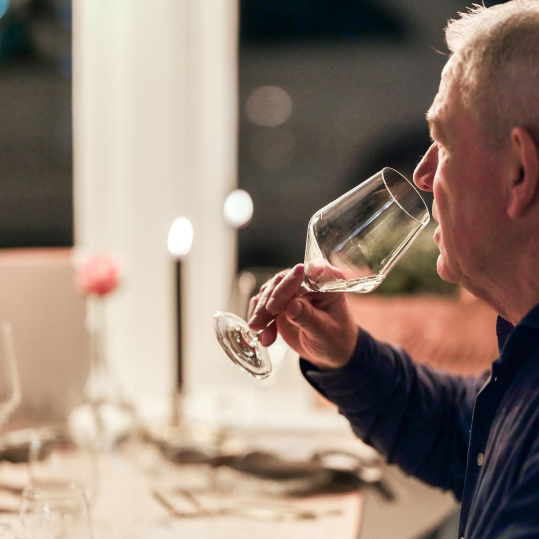 Man met grijs haar drinkt uit een wijnglas aan een sfeervol gedekte tafel met kaarsen.