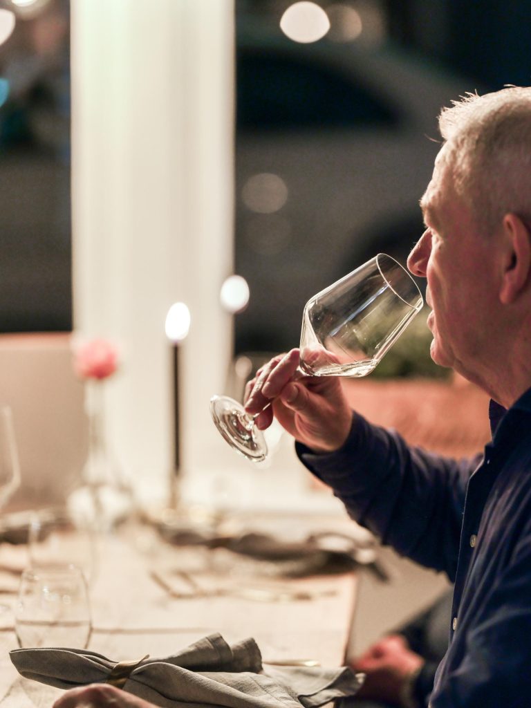 Man met grijs haar drinkt uit een wijnglas aan een sfeervol gedekte tafel met kaarsen.