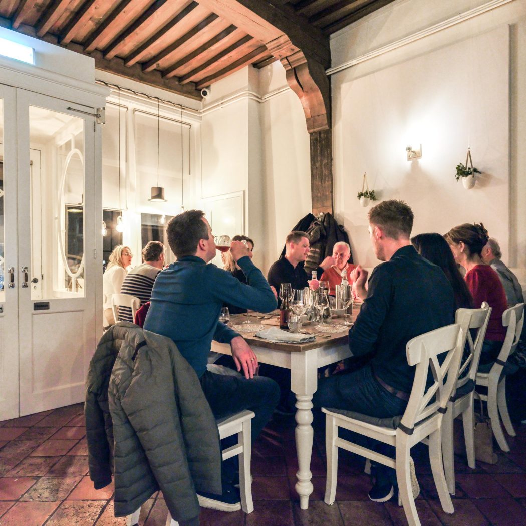 Mensen drinken wijn en eten aan een lange houten tafel in een sfeervol restaurant met houten balken en witte deuren.