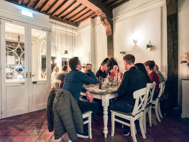 Mensen drinken wijn en eten aan een lange houten tafel in een sfeervol restaurant met houten balken en witte deuren.