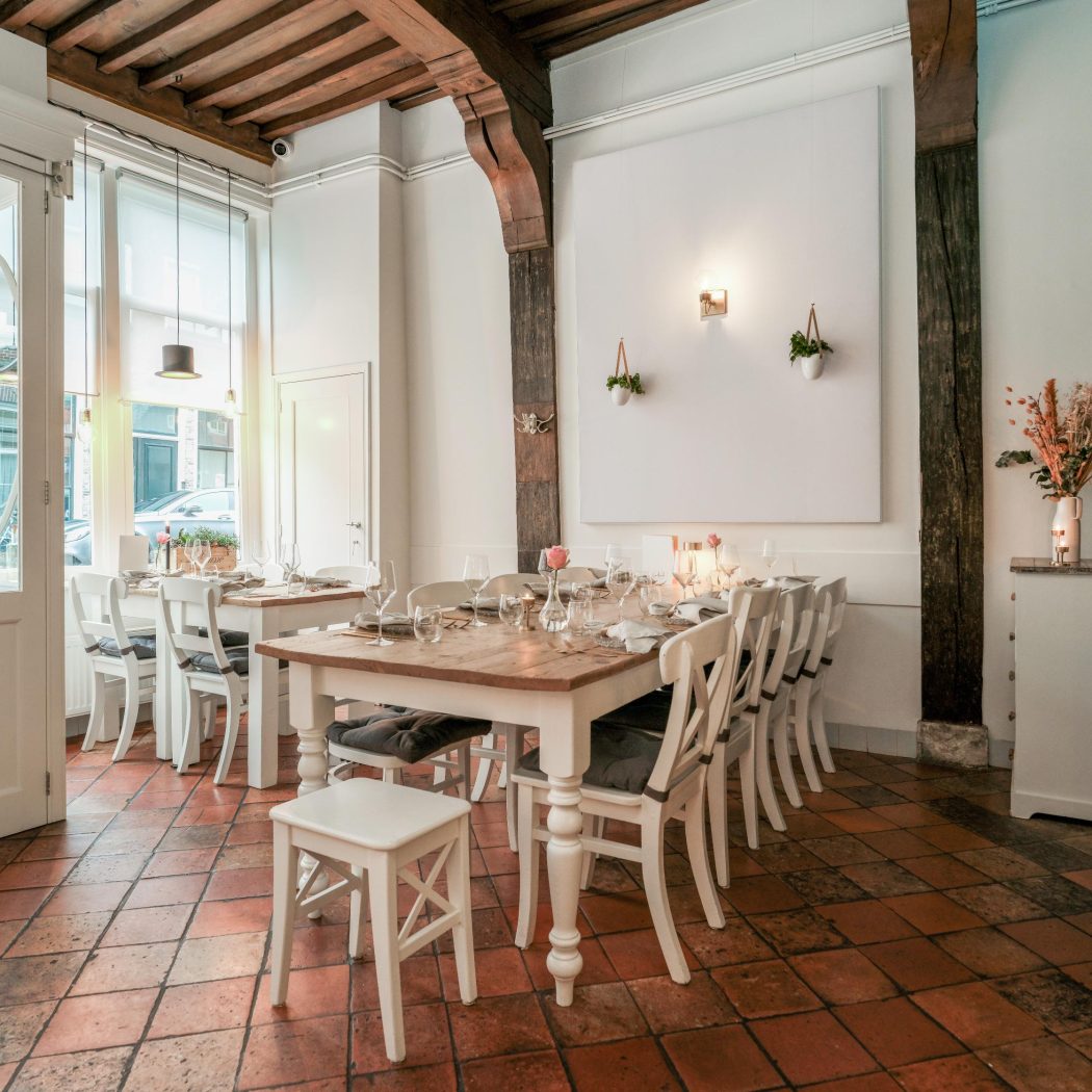 Sfeervol restaurantinterieur met houten tafels, witte stoelen, terracotta tegelvloer en TREKKEN op de glazen deur.