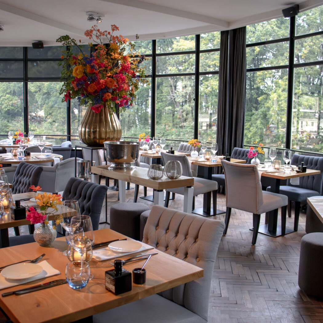 Elegant restaurantinterieur met gedekte tafels, comfortabele stoelen, een groot bloemstuk en uitzicht op groen.