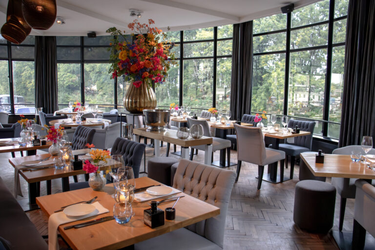 Elegant restaurantinterieur met gedekte tafels, comfortabele stoelen, een groot bloemstuk en uitzicht op groen.