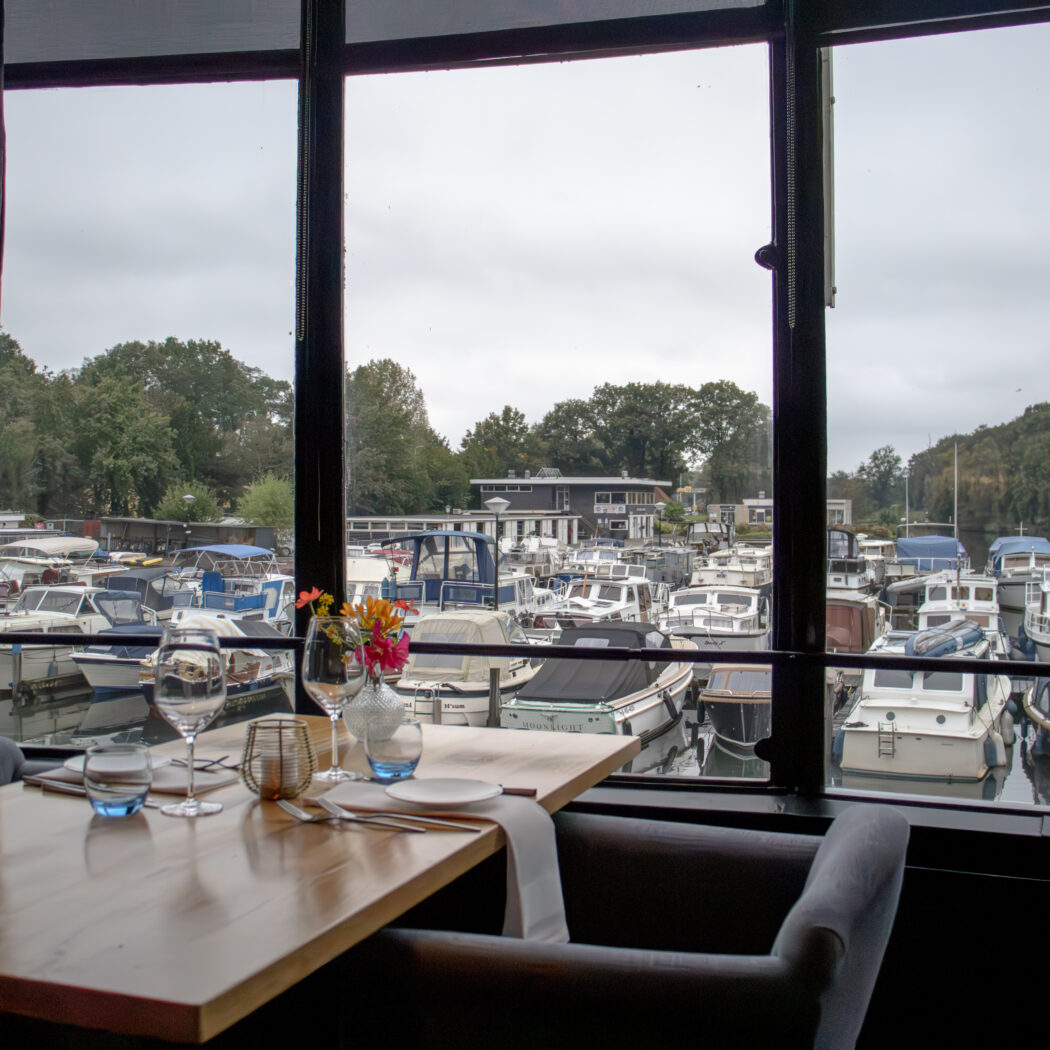 Gedekte tafel met bloemen en glazen in een restaurant, uitkijkend op een jachthaven met vele boten.