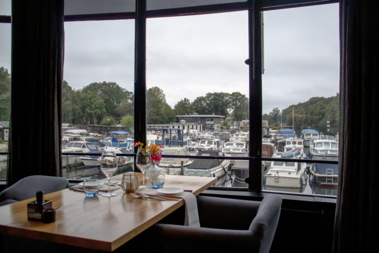 Gedekte tafel met bloemen en glazen in een restaurant, uitkijkend op een jachthaven met vele boten.