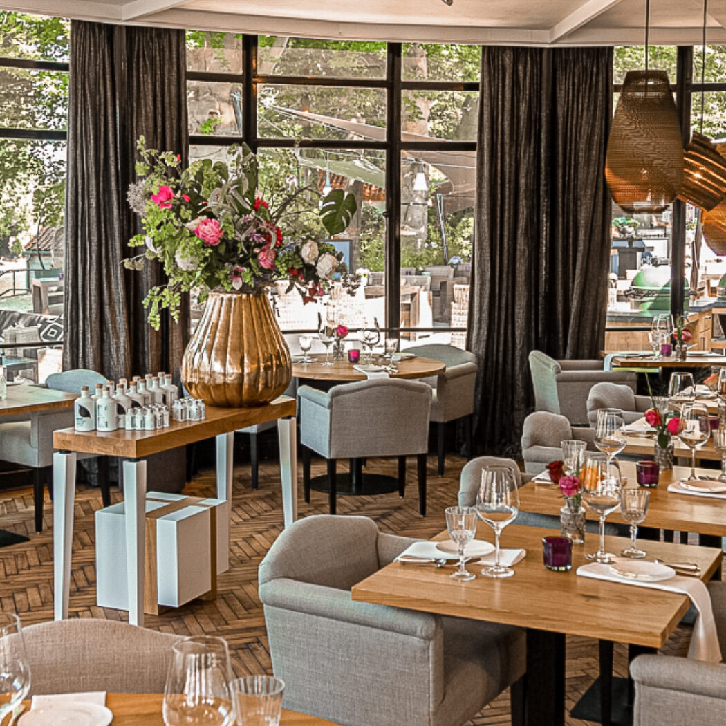 Elegant restaurant interieur met houten visgraatvloer, gedekte tafels, bloemstuk en grote ramen met terrasuitzicht.