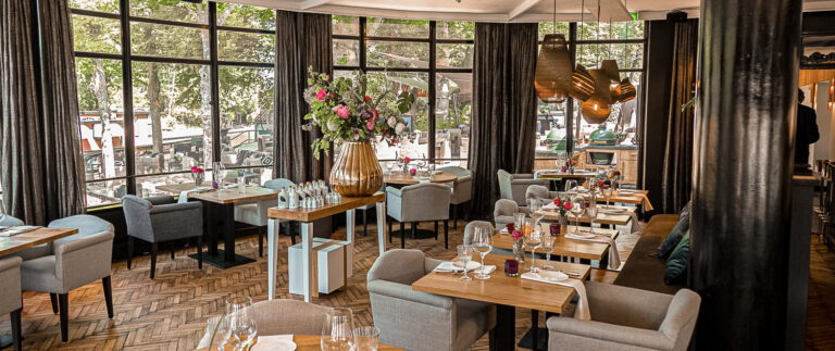 Elegant restaurant interieur met houten visgraatvloer, gedekte tafels, bloemstuk en grote ramen met terrasuitzicht.