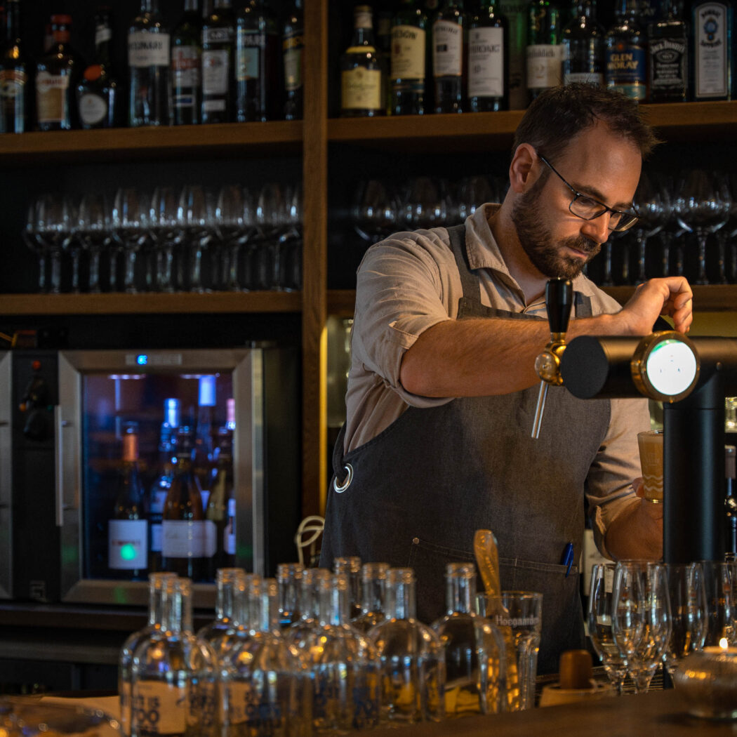 Barman met bril en schort bedient een zwarte biertap met KEIZERS display, omringd door diverse flessen en glazen.