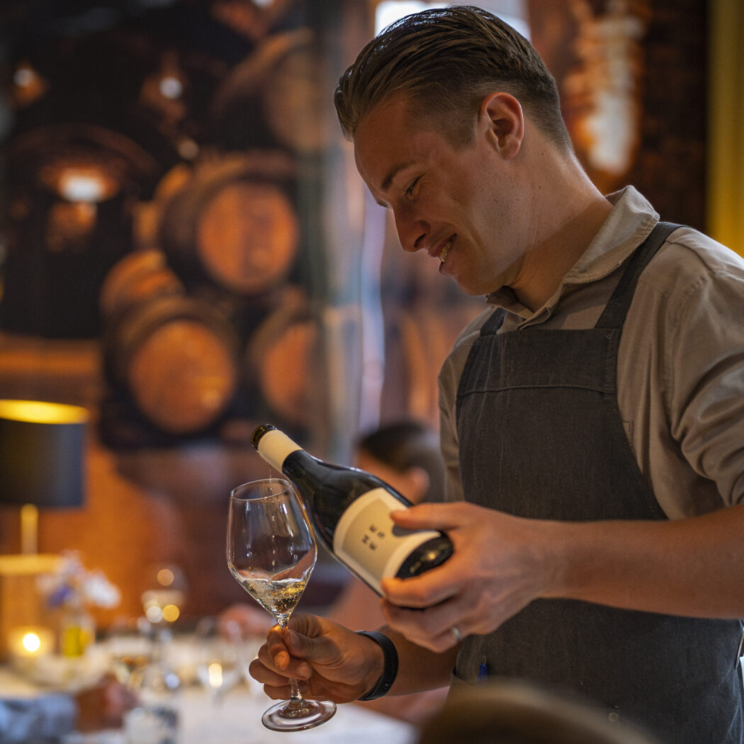 Glimlachende sommelier in grijs schort schenkt witte wijn in een glas.