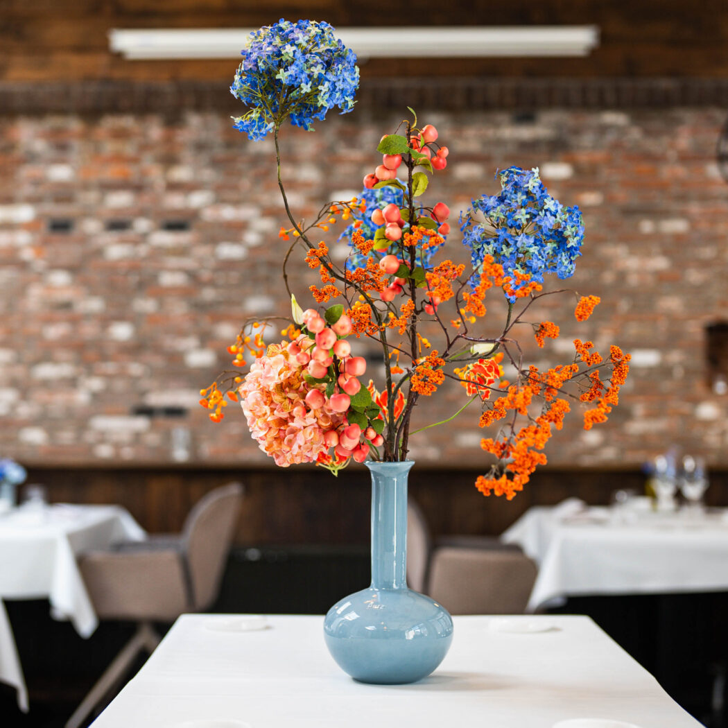 Kleurrijk bloemstuk met blauwe bloemen, perzik bessen en oranje takken in een blauwe vaas op een restauranttafel.