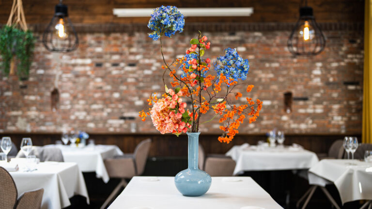 Kleurrijk bloemstuk met blauwe bloemen, perzik bessen en oranje takken in een blauwe vaas op een restauranttafel.