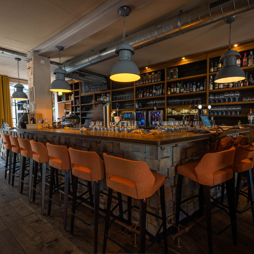 Sfeervolle bar met lange houten toonbank, oranje barkrukken, wand vol flessen en grijze hanglampen.